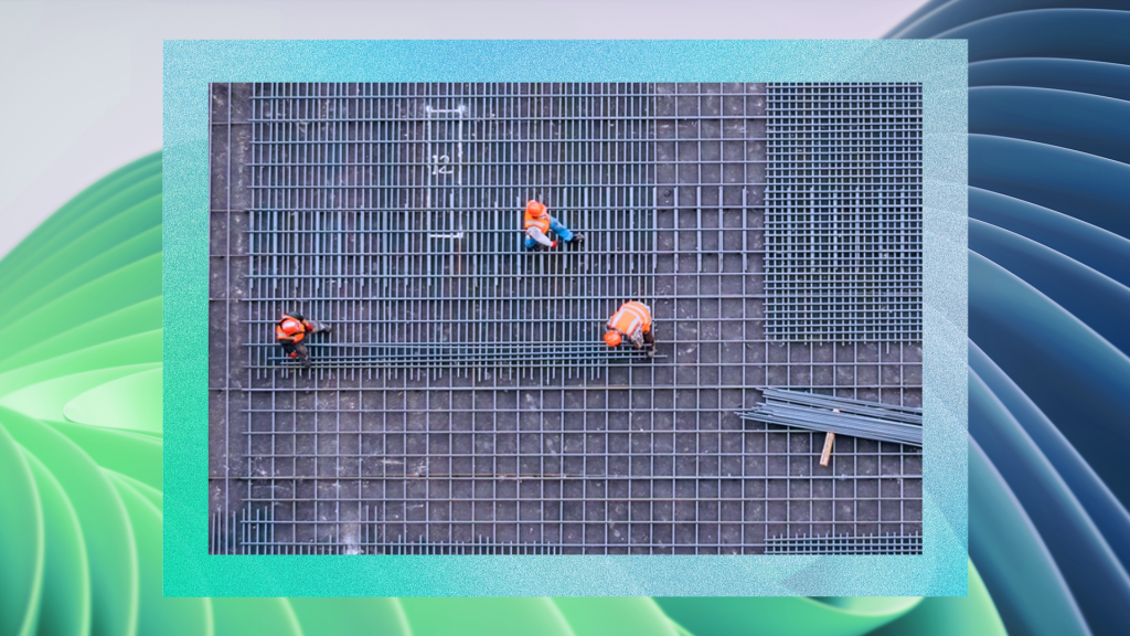Three construction workers in orange safety vests working on a large steel reinforcement grid, with metal beams placed on the right side of the structure.