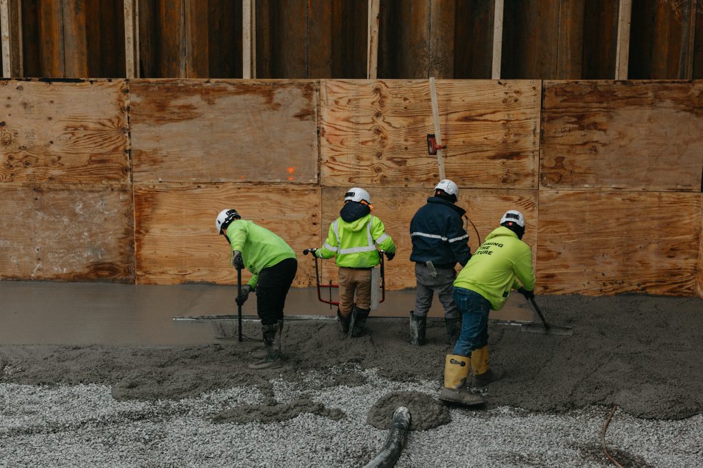 Four construction workers wearing helmets and high-visibility jackets smoothing and leveling wet concrete on a construction site, with wooden panels and a partially built wall in the background.